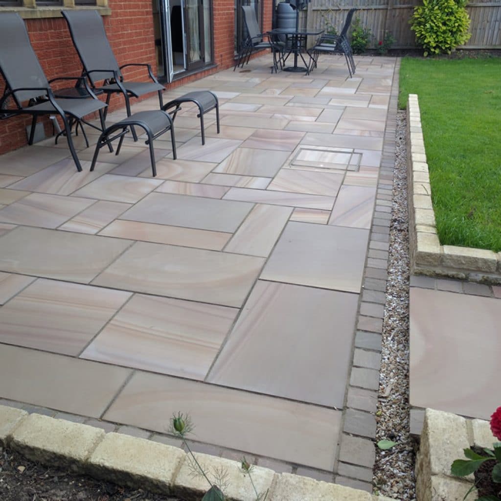 rectangular patio adjacent to brick built property. Made with rainbow sandstone and furnished with easy chairs