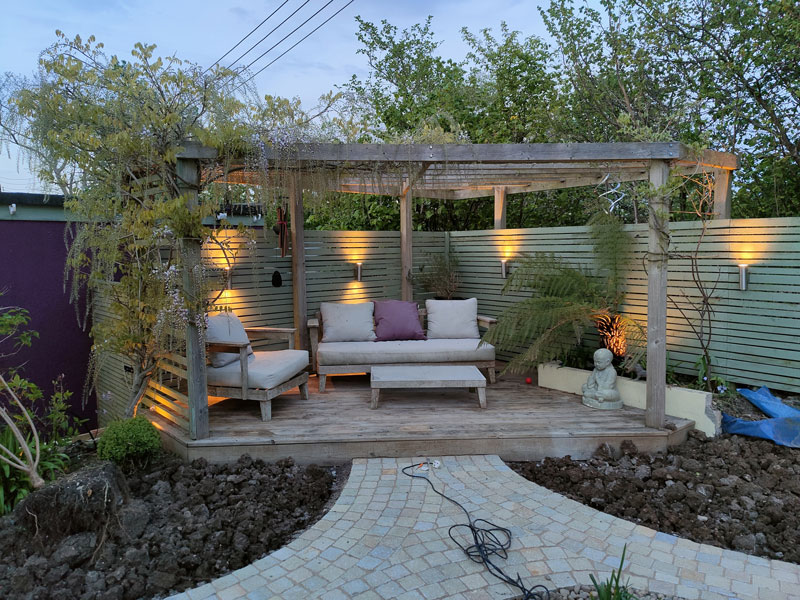 corner of a garden with bespoke decking and pergola draped with wisteria and festoon lights. 