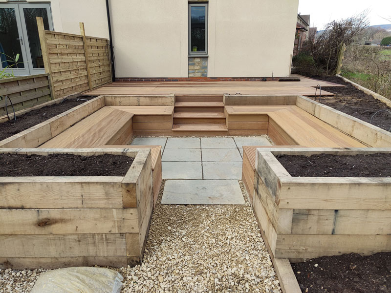 decked area beside a home with steps leading to a natural stone patio hugged by timber benches and planters